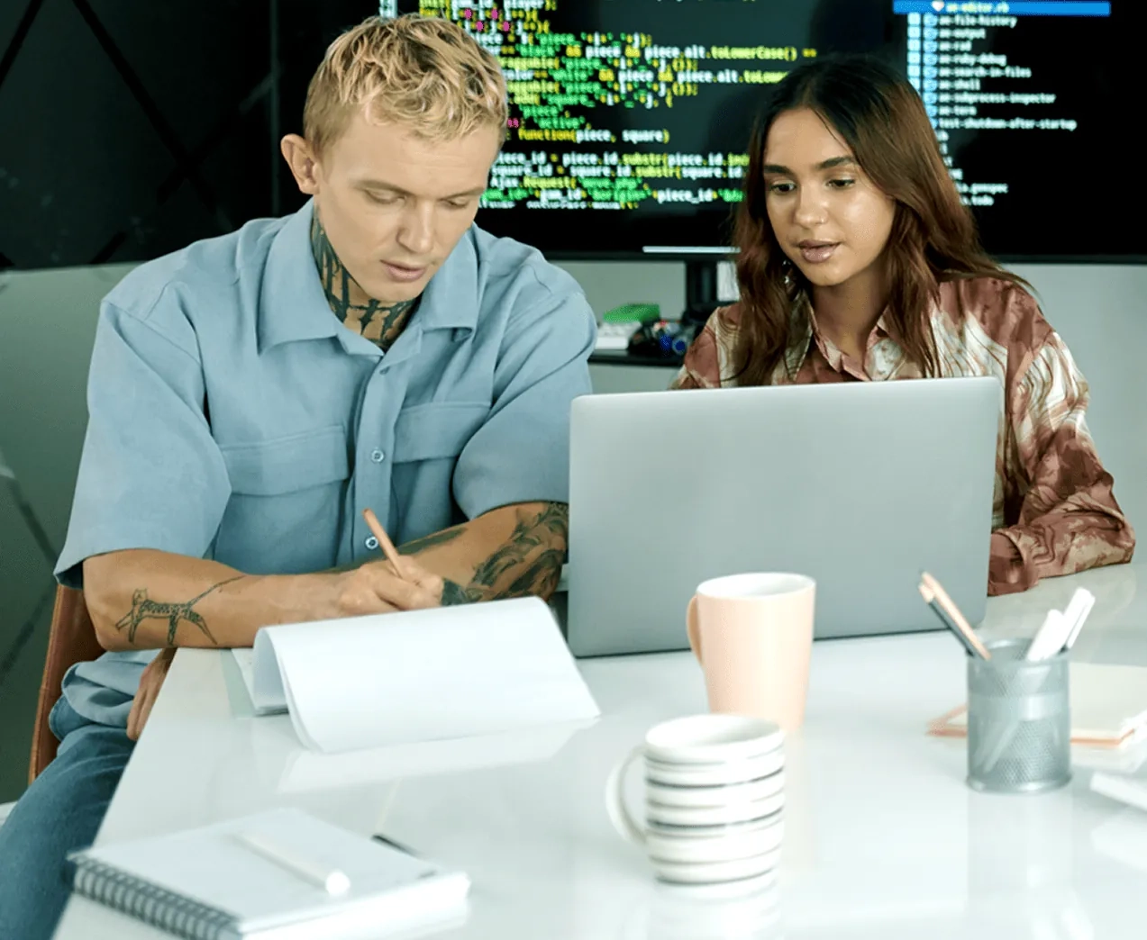 Two people look at a laptop computer in front of a screen with lines of code.