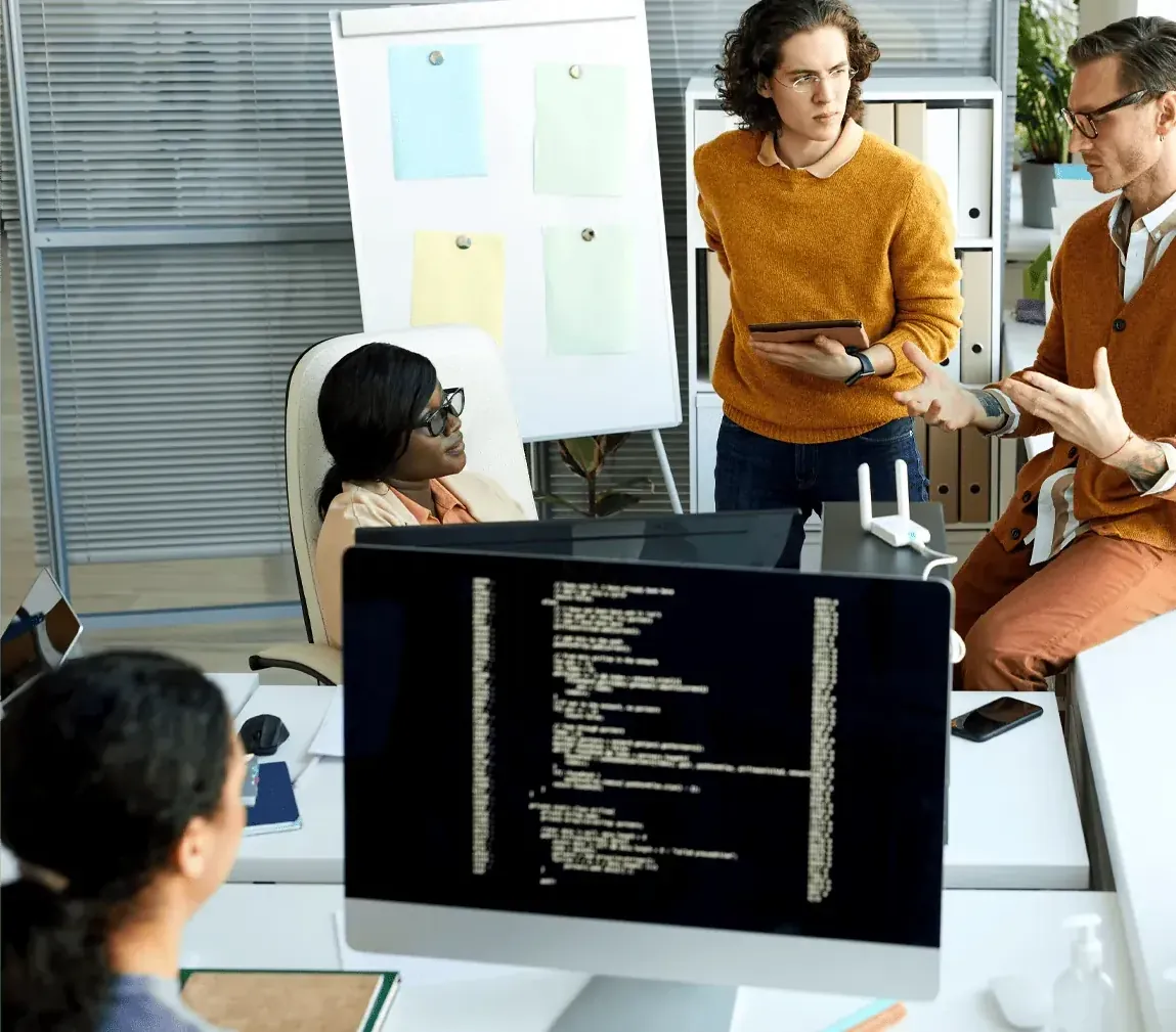 A group of people collaborate around a desk with code displayed on a monitor.